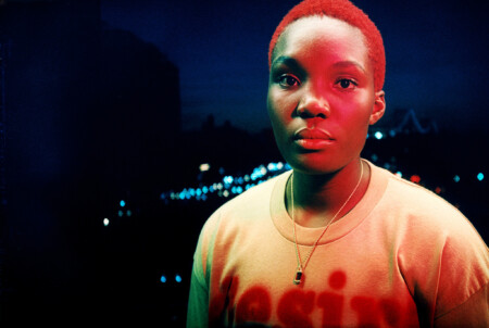 Arlo Parks steht im Dunkel draußen und wird von rot-blauen Lichtern angestrahlt.