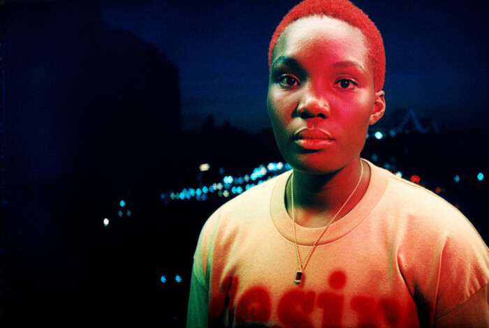 Arlo Parks steht im Dunkel draußen und wird von rot-blauen Lichtern angestrahlt.