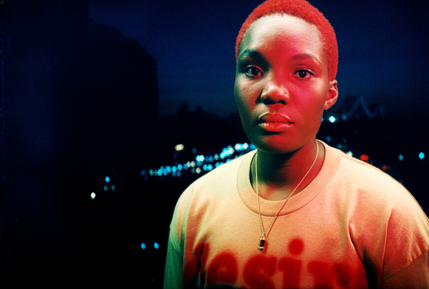 Arlo Parks steht im Dunkel draußen und wird von rot-blauen Lichtern angestrahlt.