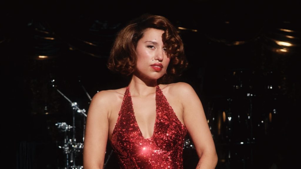 Raye steht in einem Abendkleid vor Instrumenten und schaut in die Kamera.