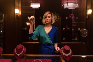 Joan steht im eleganten blau-grünen Outfit im Juweliergeschäft