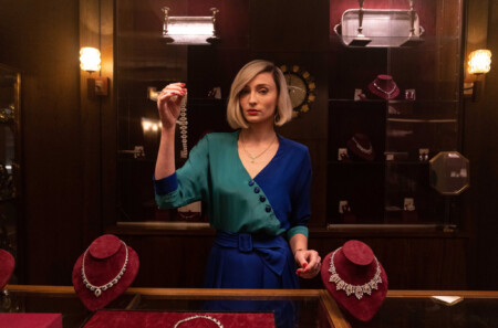 Joan steht im eleganten blau-grünen Outfit im Juweliergeschäft