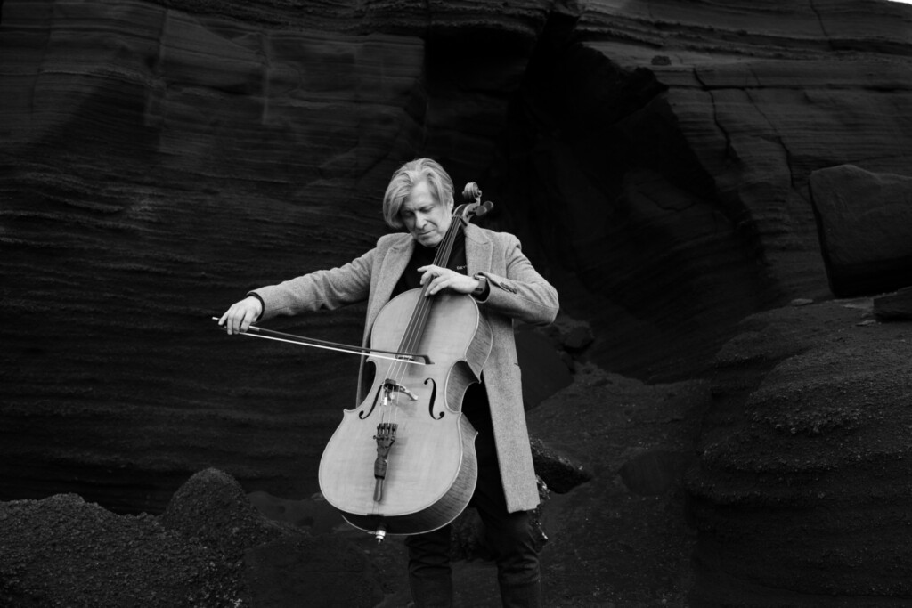 Ein schwarz-weiß Foto von Eythor Arnalds, der vor einem Felsen Cello spielt.