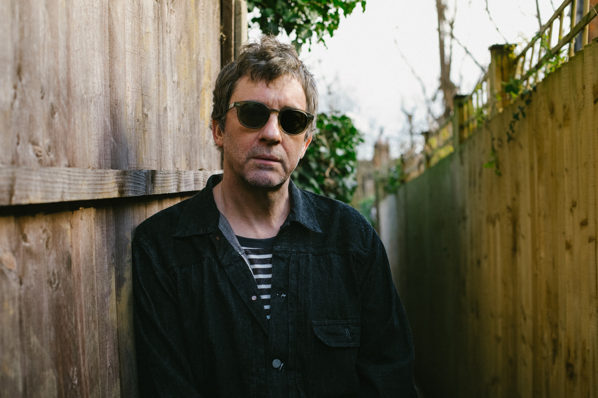 Graham Coxon steht in schwarzer Jeansjacke mit Sonnenbrille im Freien zwischen zwei Holzwänden.