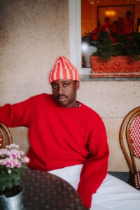 Jonny Mahoro trägt eine weiße Hose, einen roten Pulli und eine rot-weiß gestreifte Mütze. Im Hintergrund sind Stühle und Blumen auf einem Tisch zu sehen, als säße er in einem Café.