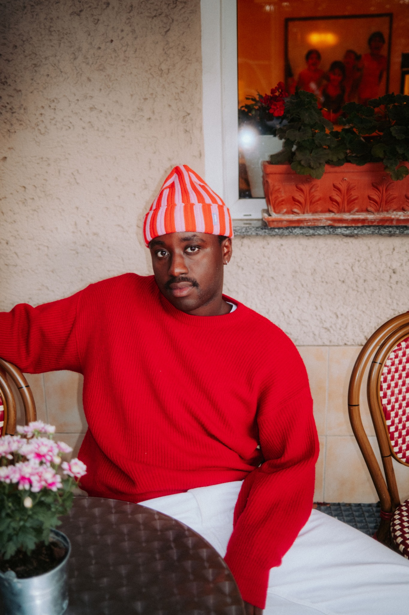 Jonny Mahoro trägt eine weiße Hose, einen roten Pulli und eine rot-weiß gestreifte Mütze. Im Hintergrund sind Stühle und Blumen auf einem Tisch zu sehen, als säße er in einem Café.