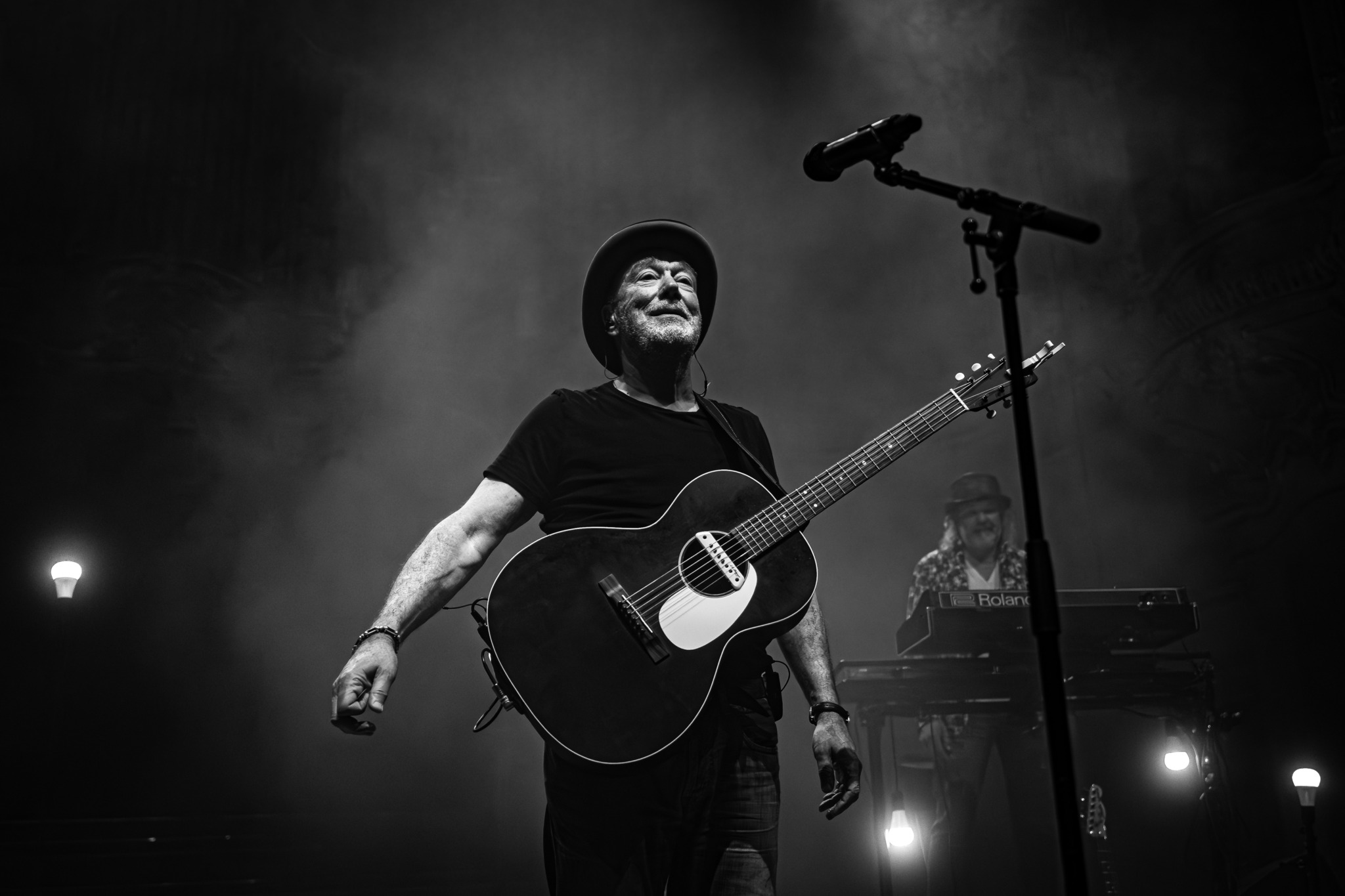 Fischer-Z Frontmann John Watts steht mit Gitarre auf einer dunklen, vernebelten Bühne.