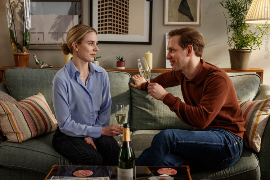 Mathilda (Carla Sehn) und Johannes (Simon Reithner) sitzen sekttrinkend auf der Couch