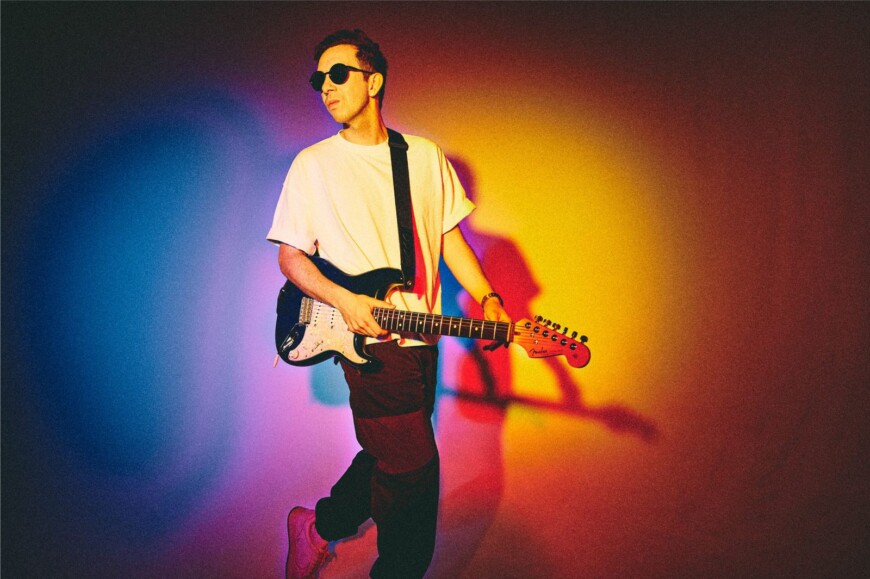Cory Wong steht mit Gitarre und Sonnebrille vor einem bunten Hintergrund