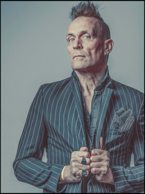 John Robb wird bei „A Life without Punk“ auftreten. Hier trägt er ein gestreiftes Jacket, das er am Revere festhält.