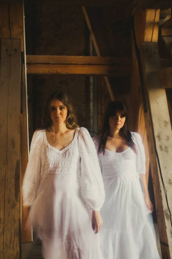 Klara und Johanna Söderberg von First Aid Kit stehen in weißen Kleidern in einem hölzernen Durchgang