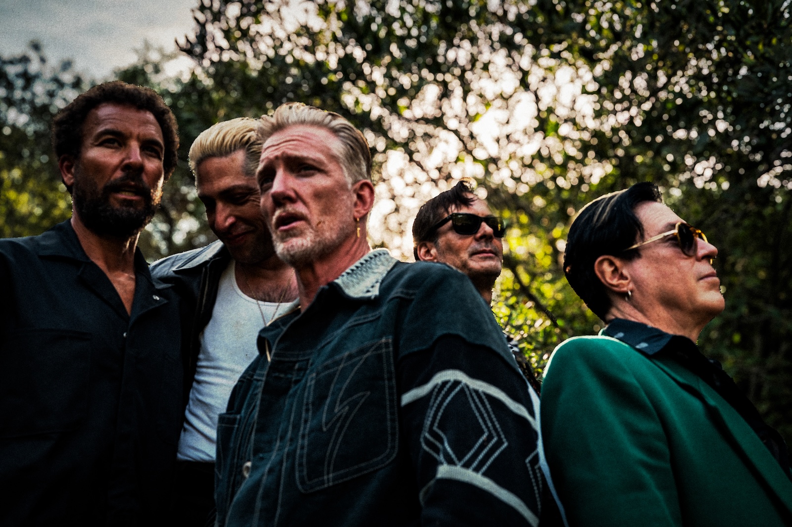 Köpfe und Oberkörper der fünf Musiker von Queens of the Stone Age. Sie wurden draußen leicht von unten fotografiert; über ihnen sind Baumwipfel zu sehen.