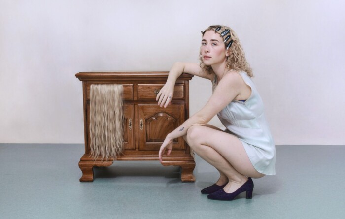 Sängerin Blondshell hockt auf dem Boden neben einem Antik-Holz Regal.