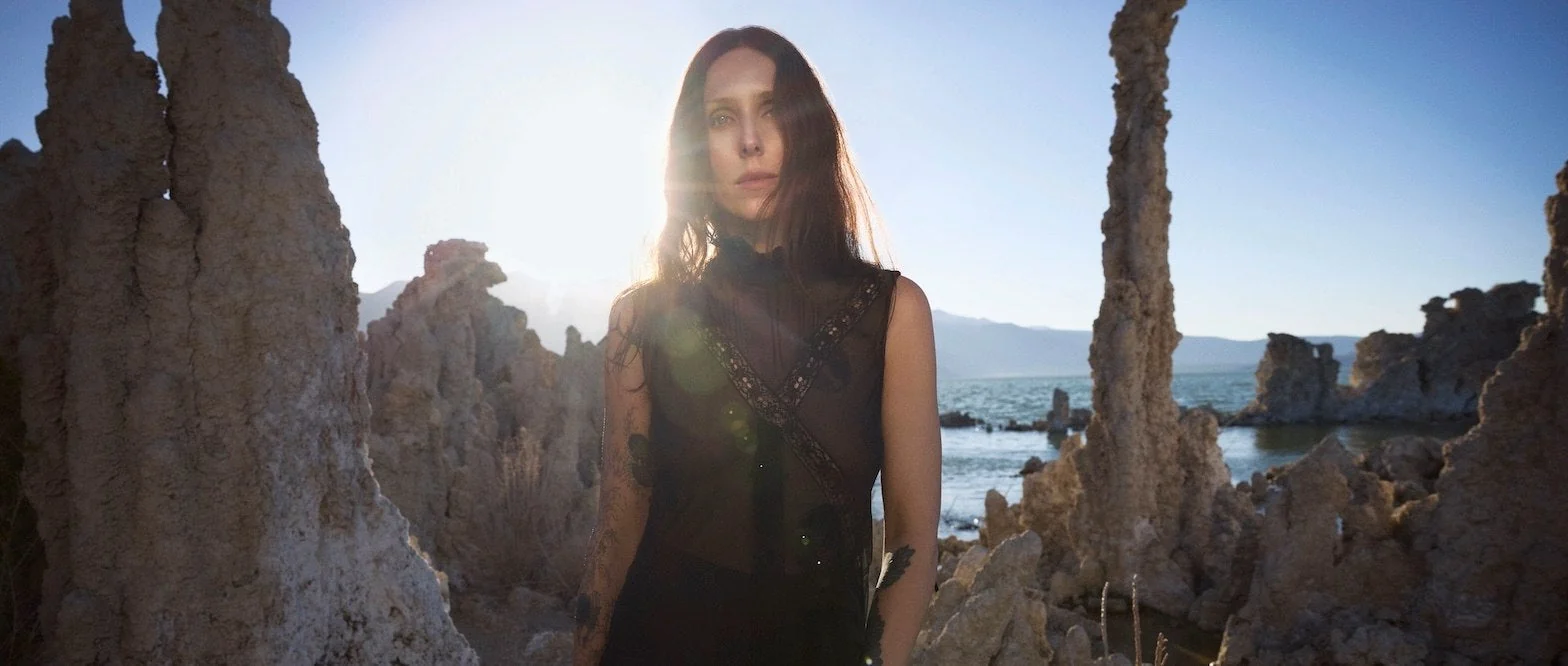 Chelsea Wolfe steht in einer Kulisse aus felsen am Meer und blickt in die Kamera.
