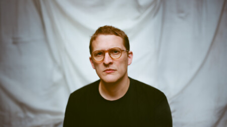 Sam Shepherd alias Floating Points sitzt vor einem weißen Vorhang in einem Raum.