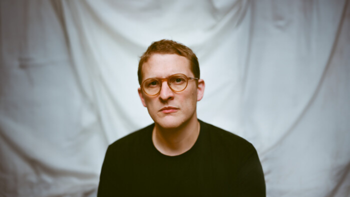 Sam Shepherd alias Floating Points sitzt vor einem weißen Vorhang in einem Raum.