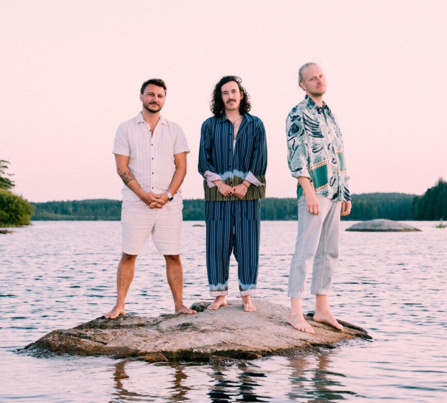 Die drei Bandmitglieder stehen auf einer Mini-Insel inmitten Wasser