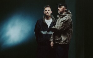 Das Musikduo Tom und Ed Russell stehen vor schwarz-blauen Hintergrund.