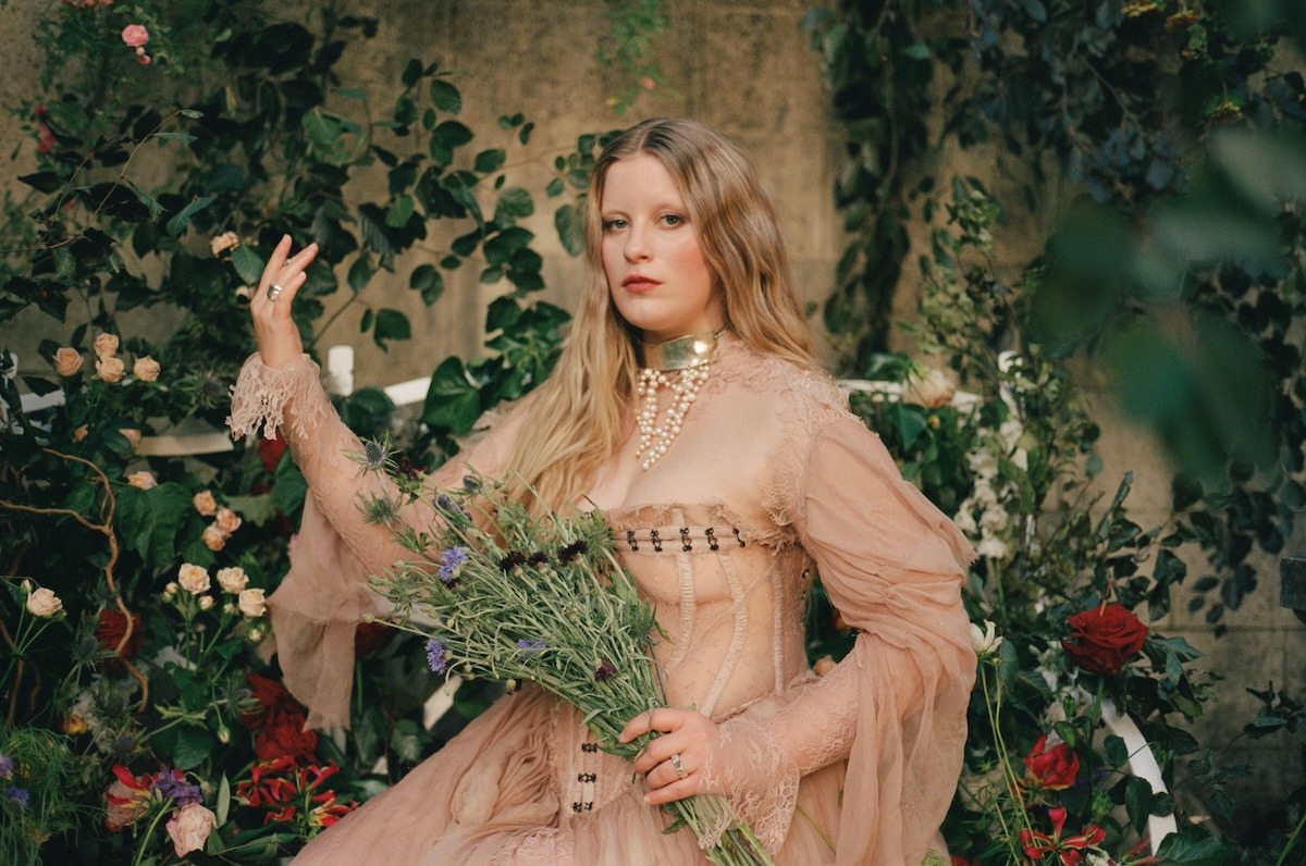 Paris Paloma sitzt in einem barocken Kleid in einem Garten voller Blumen.