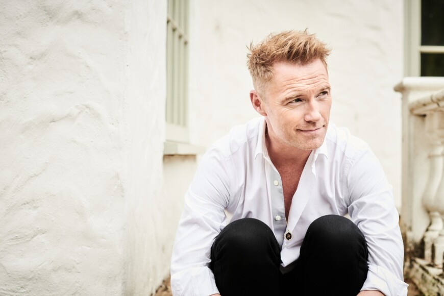Ronan Keating sitzt im weißen Hemd mit angezogenen Beinen vor einer Hauswand
